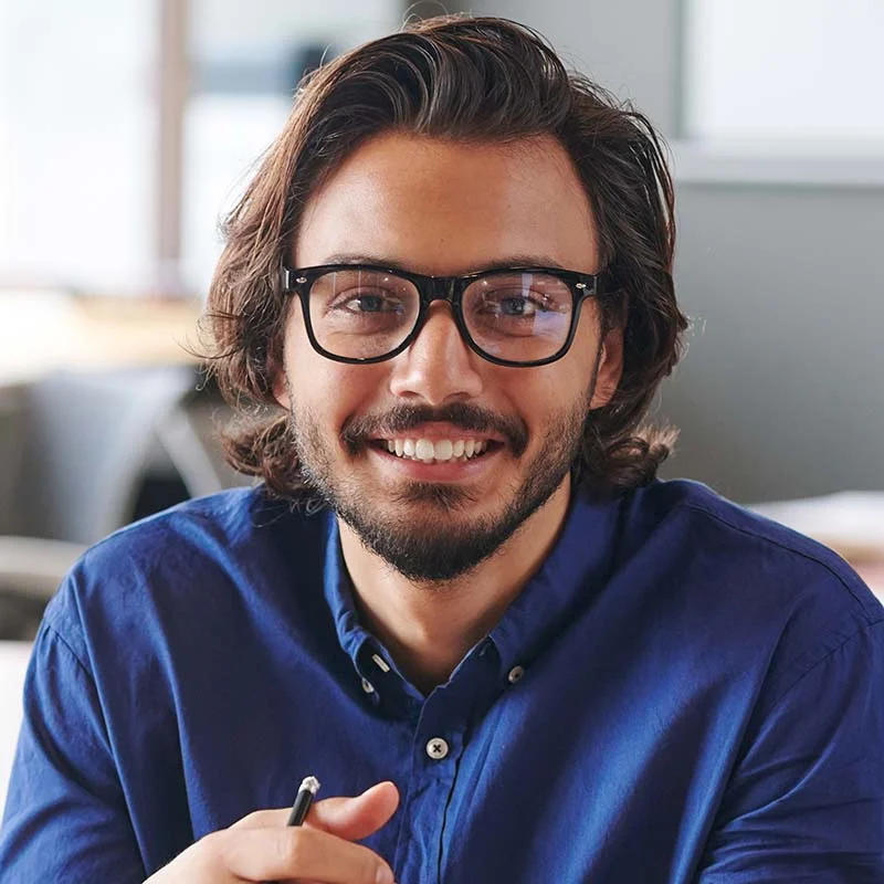 Um homem com cabelos castanhos na altura dos ombros e barba sorri calorosamente para a câmera. Ele está usando óculos de aros pretos e uma camisa azul de botões. O fundo parece ser um ambiente interno com elementos desfocados, possivelmente um escritório ou espaço de estudo. Ele está segurando uma caneta na mão direita. | Morabeza Marketing