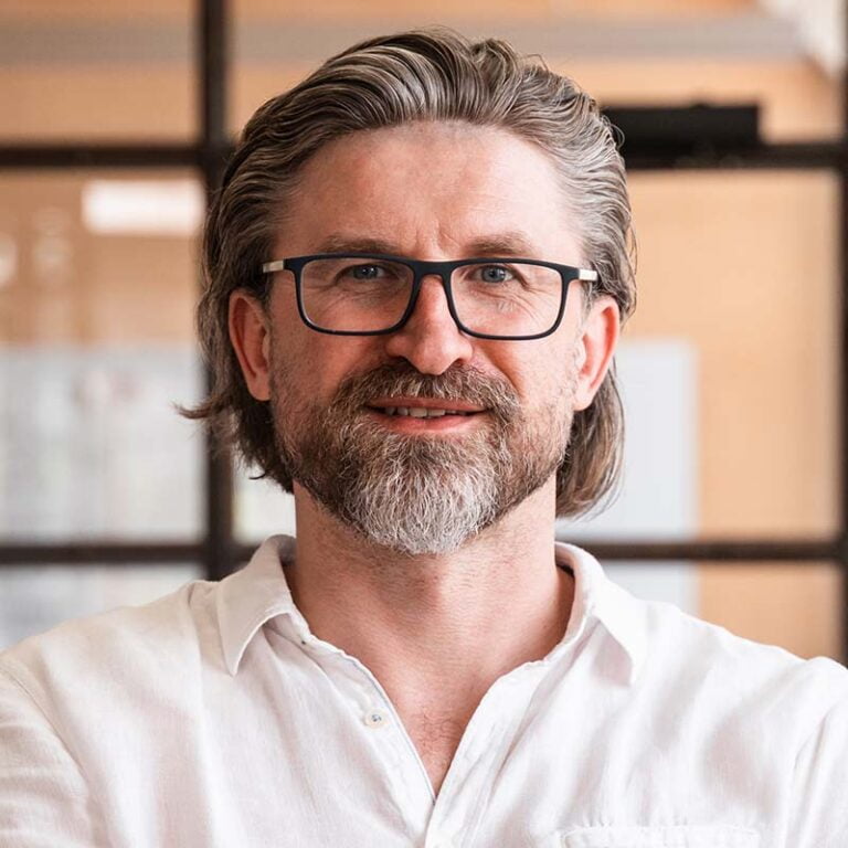 A man with shoulder-length grey hair and a full beard, wearing black-framed glasses and a white button-down shirt, stands against a blurred interior background of glass doors and beige walls. He is smiling slightly and looks confident and approachable. | Morabeza Marketing