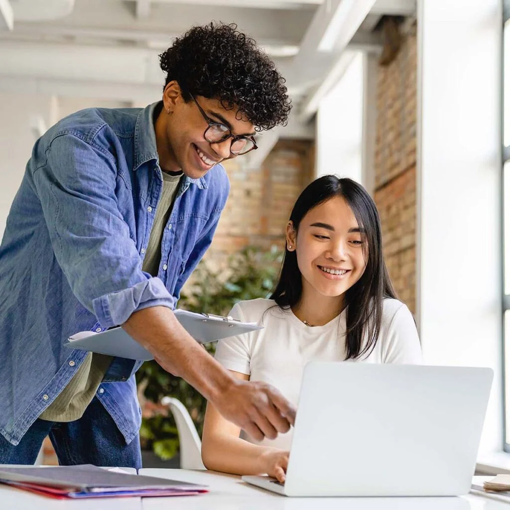 Em um ambiente de escritório bem iluminado, um homem sorridente com cabelos cacheados, usando óculos e uma camisa jeans, se inclina para ajudar uma mulher com longos cabelos escuros, que está sorrindo para a tela de um laptop. A mulher está sentada e vestindo uma camisa branca. Eles parecem estar colaborando em um projeto. | Morabeza Marketing