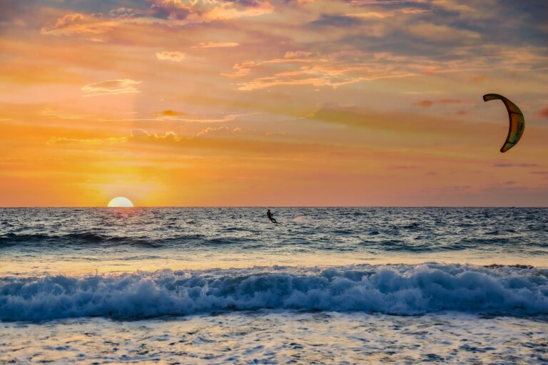 A person parasailing over the ocean during a vibrant sunset in Cape Verde. The sun is partially visible just above the horizon, casting an orange and pink glow in the sky and reflecting on the water. The waves gently lap the shore in the foreground, creating a serene atmosphere. | Morabeza Marketing