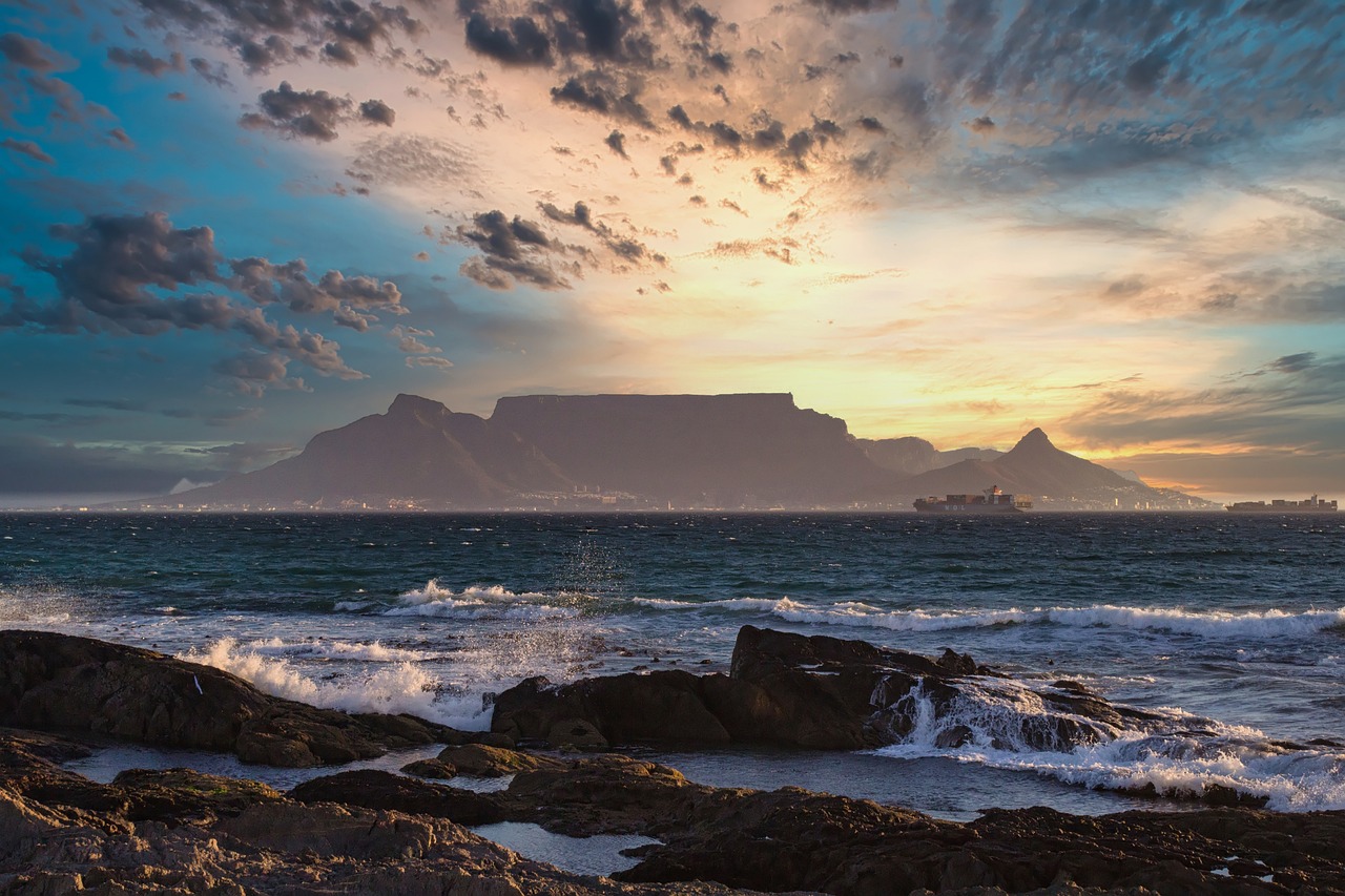 Pôr do sol sobre a Table Mountain na Cidade do Cabo, África do Sul, com a silhueta da montanha contra um céu vibrante cheio de nuvens de várias cores. O primeiro plano apresenta ondas quebrando em costas rochosas, uma reminiscência de estratégias de SEO para os pontos críticos costeiros de Cabo Verde, e um navio pode ser visto à distância no oceano. | Morabeza Marketing